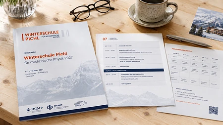 Druckunterlagen für die Winterschule Pichl 2027, eine Brille, ein Stift und eine Kaffeetasse auf einem Holztisch.